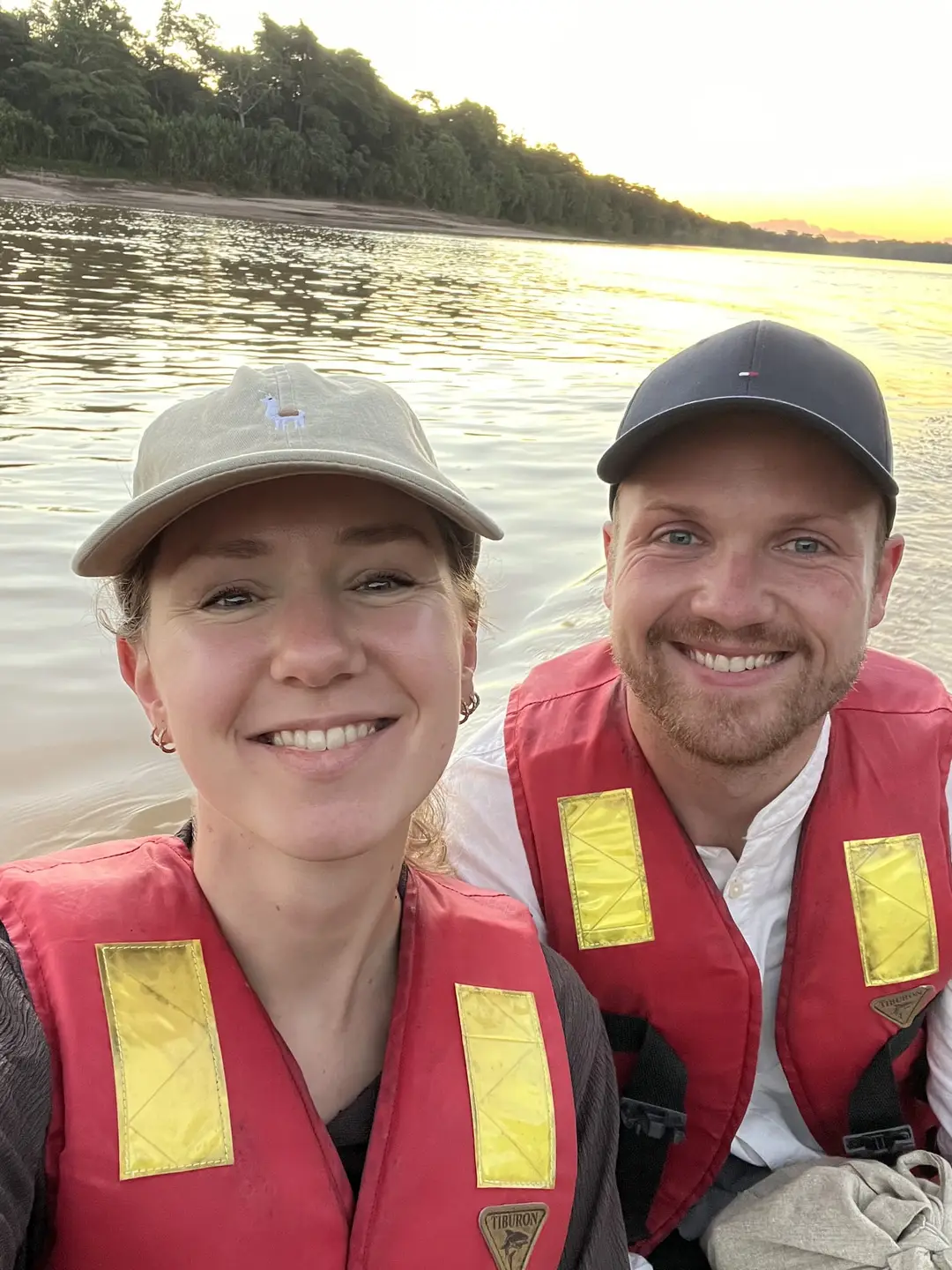 Selfie von Rebecca und Max in roten Schwimmwesten im Boot, hinter ihnen der Fluss und bewaldetes Ufer im Abendlicht