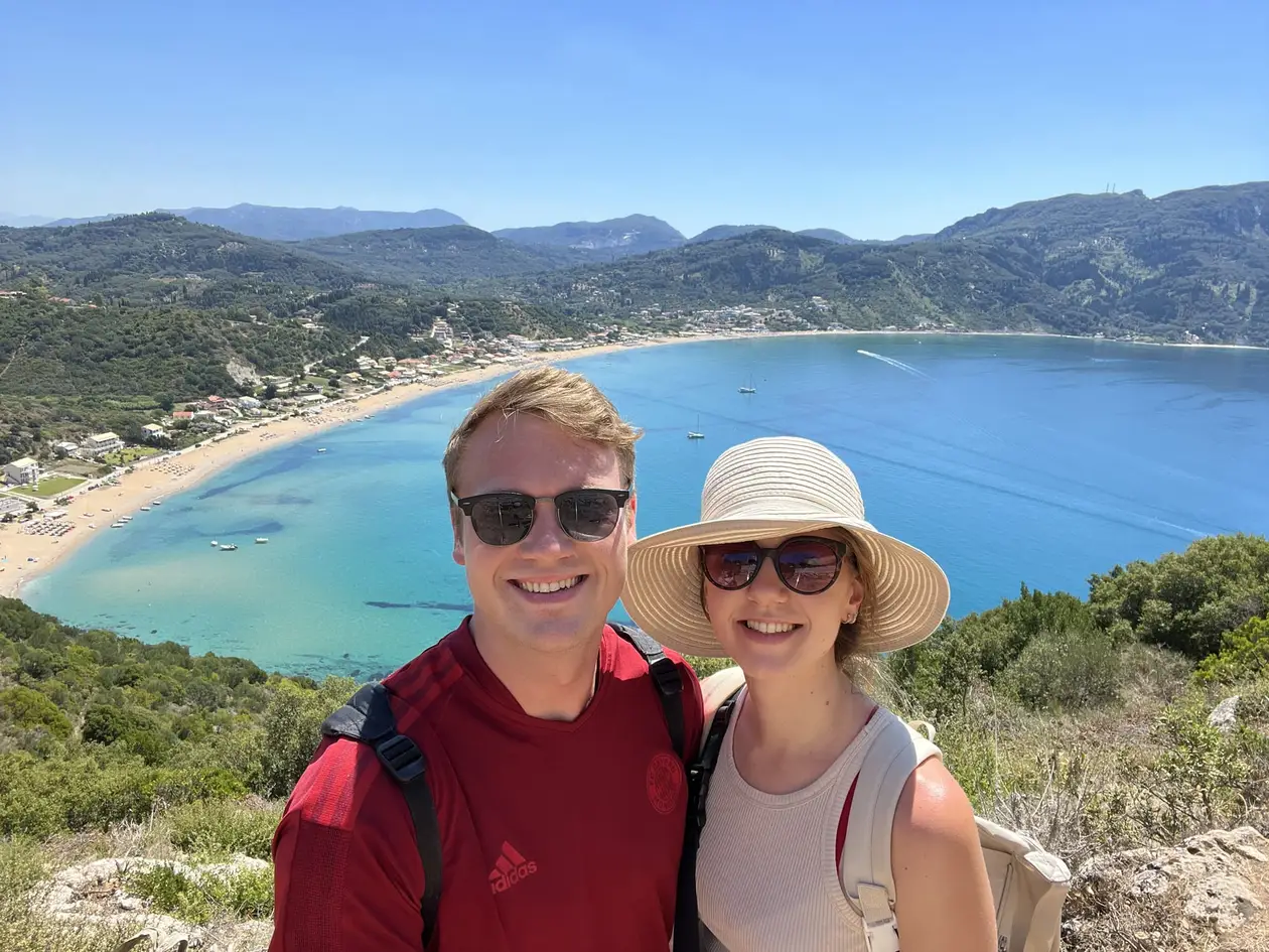 Selfie von Max und Rebecca mit Sonnenhut, hinter ihnen tief unten eine weite Bucht mit langem Sandstrand und Bergen