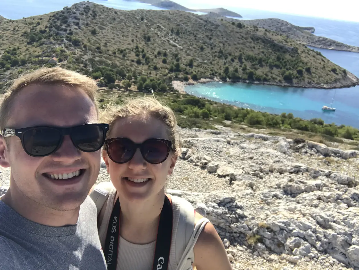 Selfie von Max und Rebecca mit Sonnenbrillen auf felsigem Kalkboden, hinter ihnen eine kahle Insel mit türkiser Bucht und einem Segelboot
