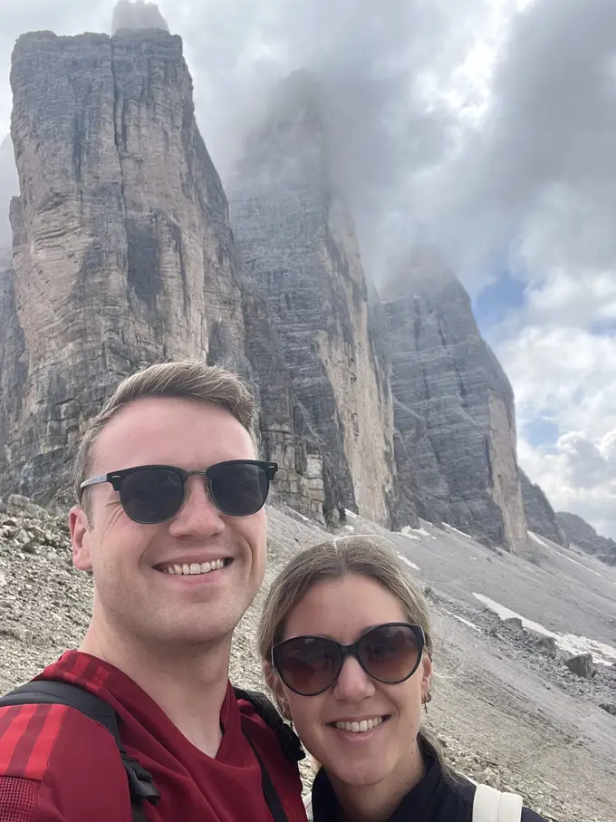 Selfie von Max und Rebecca mit Sonnenbrillen, hinter ihnen ragen die Felswände der Drei Zinnen in die Wolken