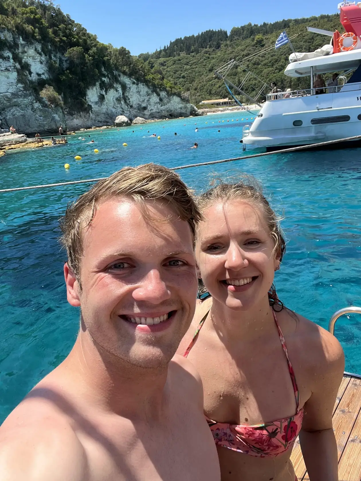 Selfie von Max und Rebecca mit nassen Haaren auf einem Boot, hinter ihnen türkises Wasser und eine Motoryacht