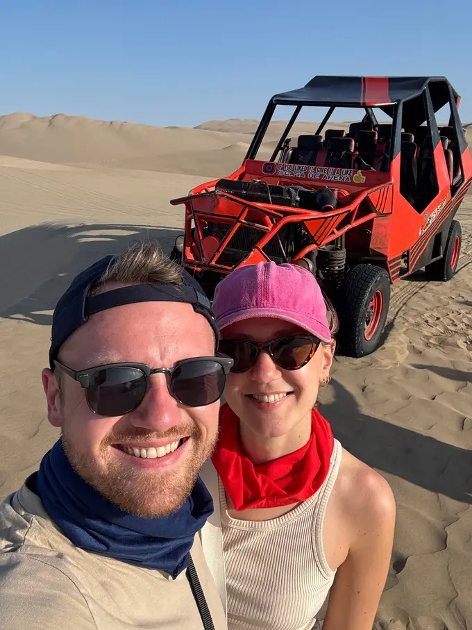 Max und Rebecca mit Tüchern um den Hals und Sonnenbrillen, hinter ihnen ein roter Buggy auf den Dünen