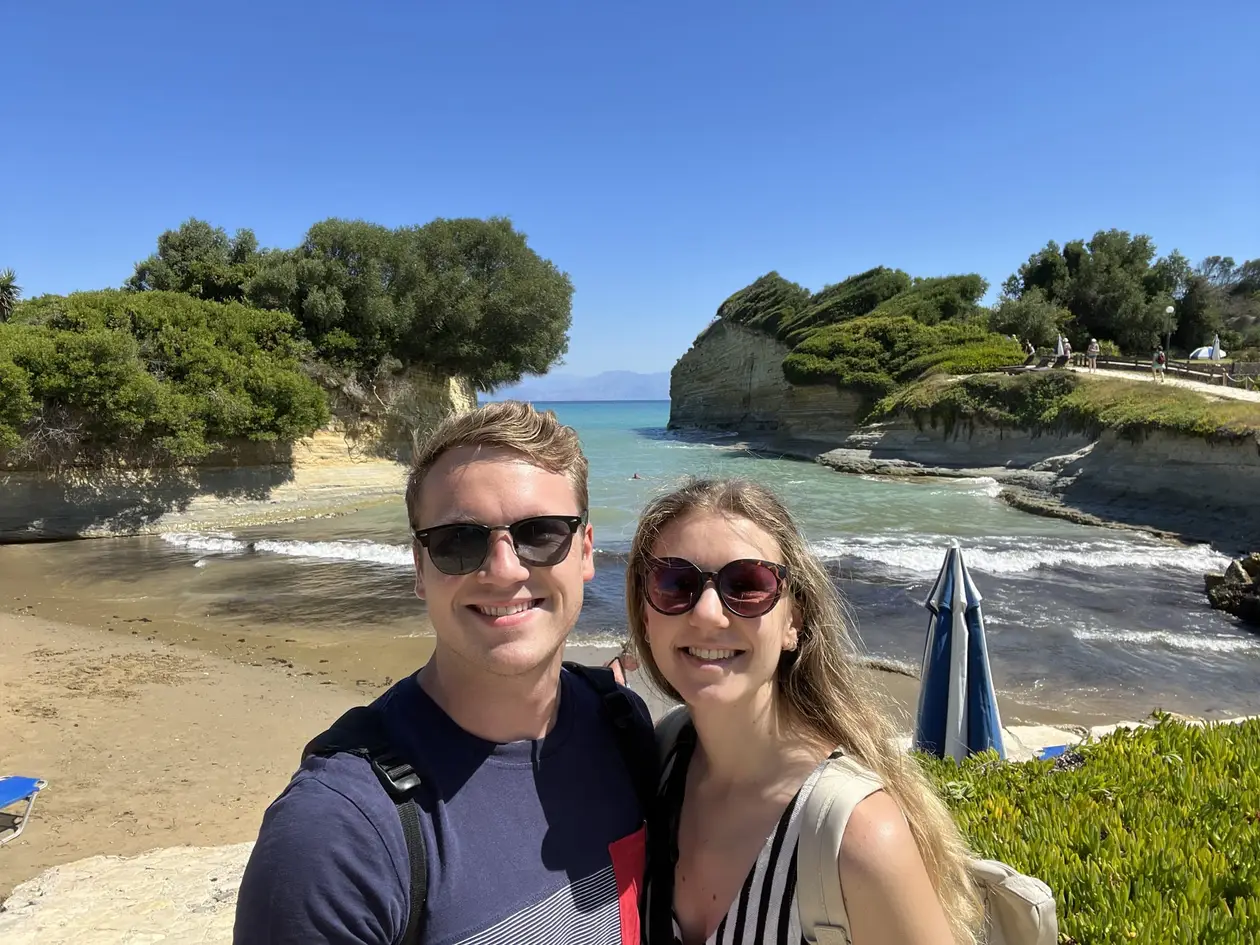 Selfie von Max und Rebecca mit Sonnenbrillen, hinter ihnen zwei bewachsene Sandsteinfelsen und eine Bucht