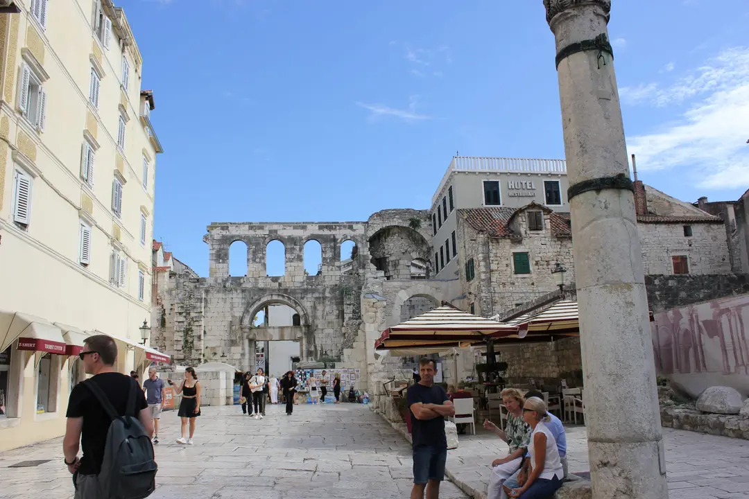 Platz vor einem Tor des Diokletianpalasts mit Bogenreihe und Arkaden, davor Passanten, ein Café und eine antike Säule