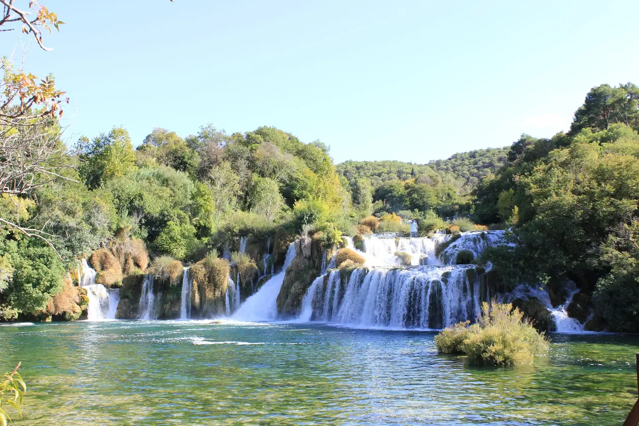 Die breite Kaskade von Skradinski Buk, das Wasser fällt über mehrere bewachsene Sinterstufen in ein grünes Becken, ringsum Wald