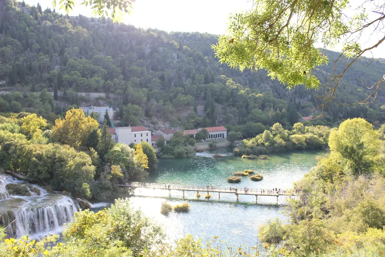 Blick durch Laub von oben auf die Kaskade von Skradinski Buk, rechts eine lange Holzbrücke mit Besuchern über dem Fluss, dahinter Steinhäuser mit roten Dächern am bewaldeten Hang