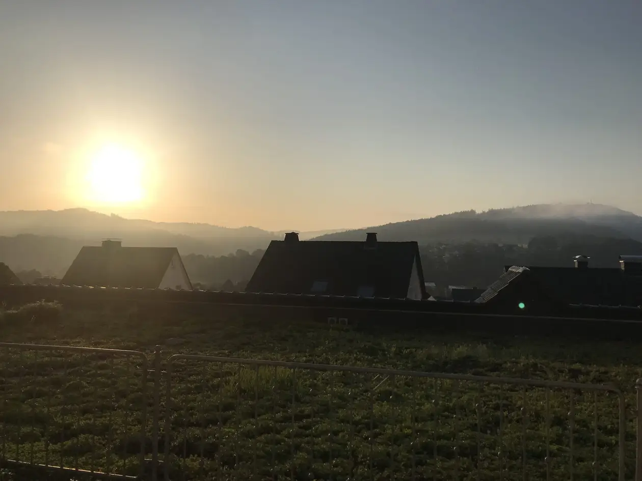 Sonnenaufgang über bewaldeten Hügeln mit Morgendunst, im Vordergrund ein begrüntes Flachdach, ein Bauzaun und die dunklen Dächer der Nachbarhäuser
