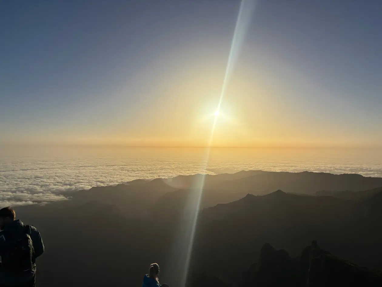 Weites Wolkenmeer zwischen schroffen Gipfeln im Morgenlicht, die Sonne steht tief über dem Horizont
