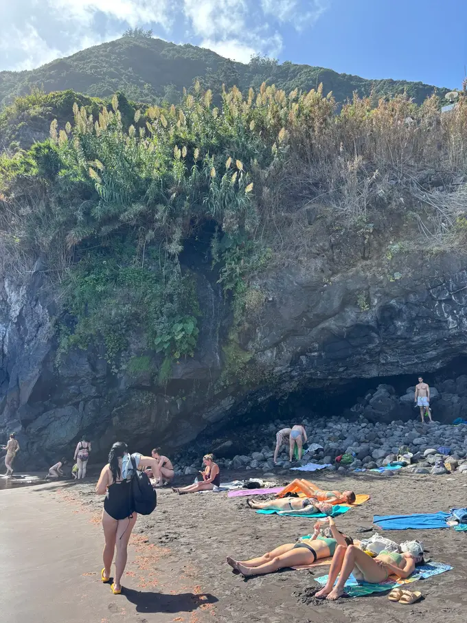 Mehrere Personen liegen auf Handtüchern auf schwarzem Sand, dahinter ein steiler grüner Hang
