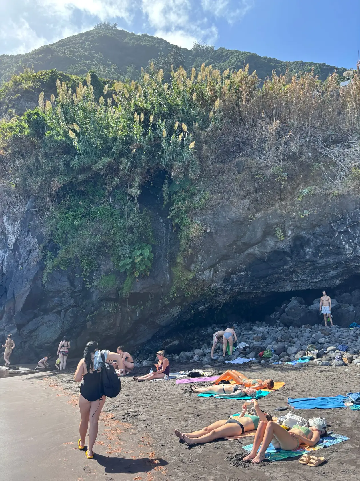Mehrere Personen liegen auf Handtüchern auf schwarzem Sand, dahinter ein steiler grüner Hang