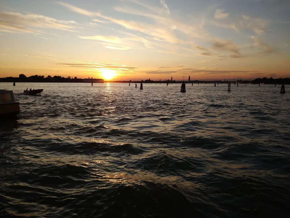 Die Sonne geht über der Lagune unter, im Vordergrund dunkles Wasser mit Pfählen und einem Boot