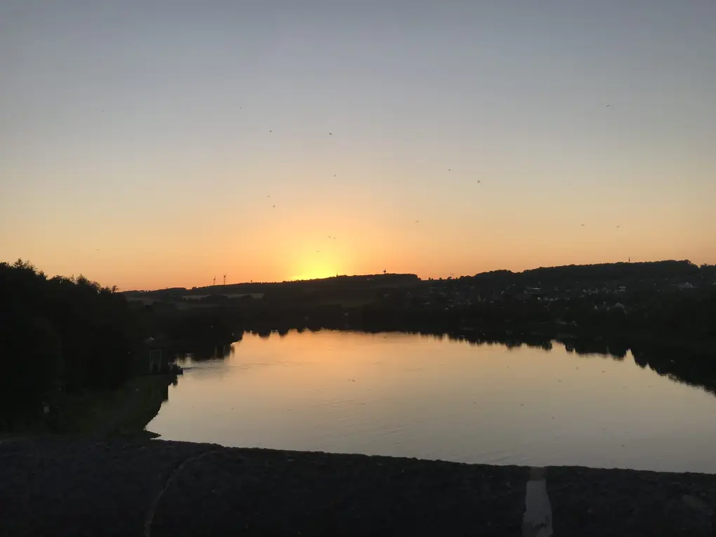 Die Sonne ist gerade hinter einer Hügelkette mit Windrädern untergegangen, der Himmel ist orange, darunter spiegelt sich ein Gewässer im Tal, vorne die dunkle Brüstung der Staumauer