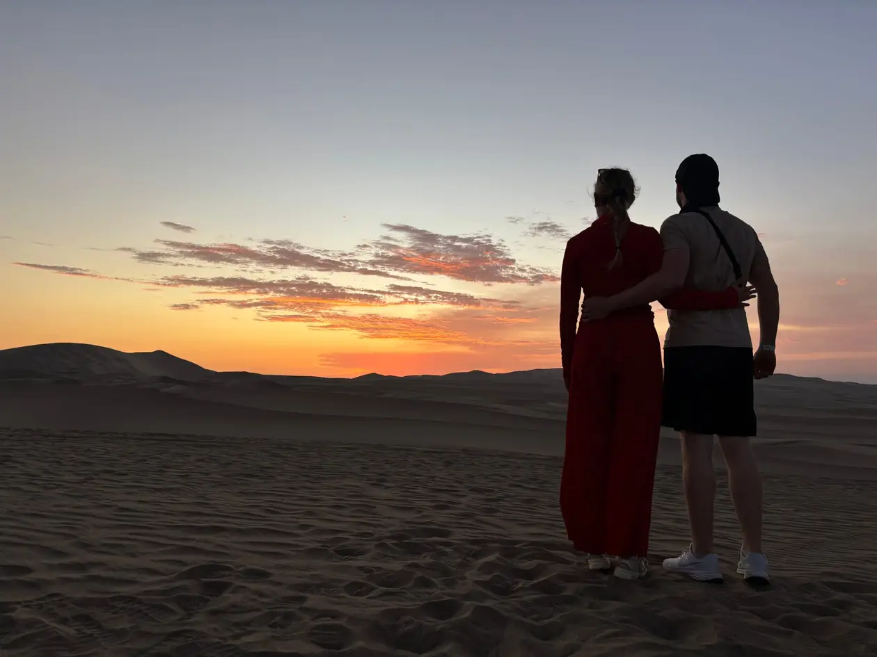 Max und Rebecca stehen als Silhouette Arm in Arm auf einer Düne und schauen in den orangen Sonnenuntergang