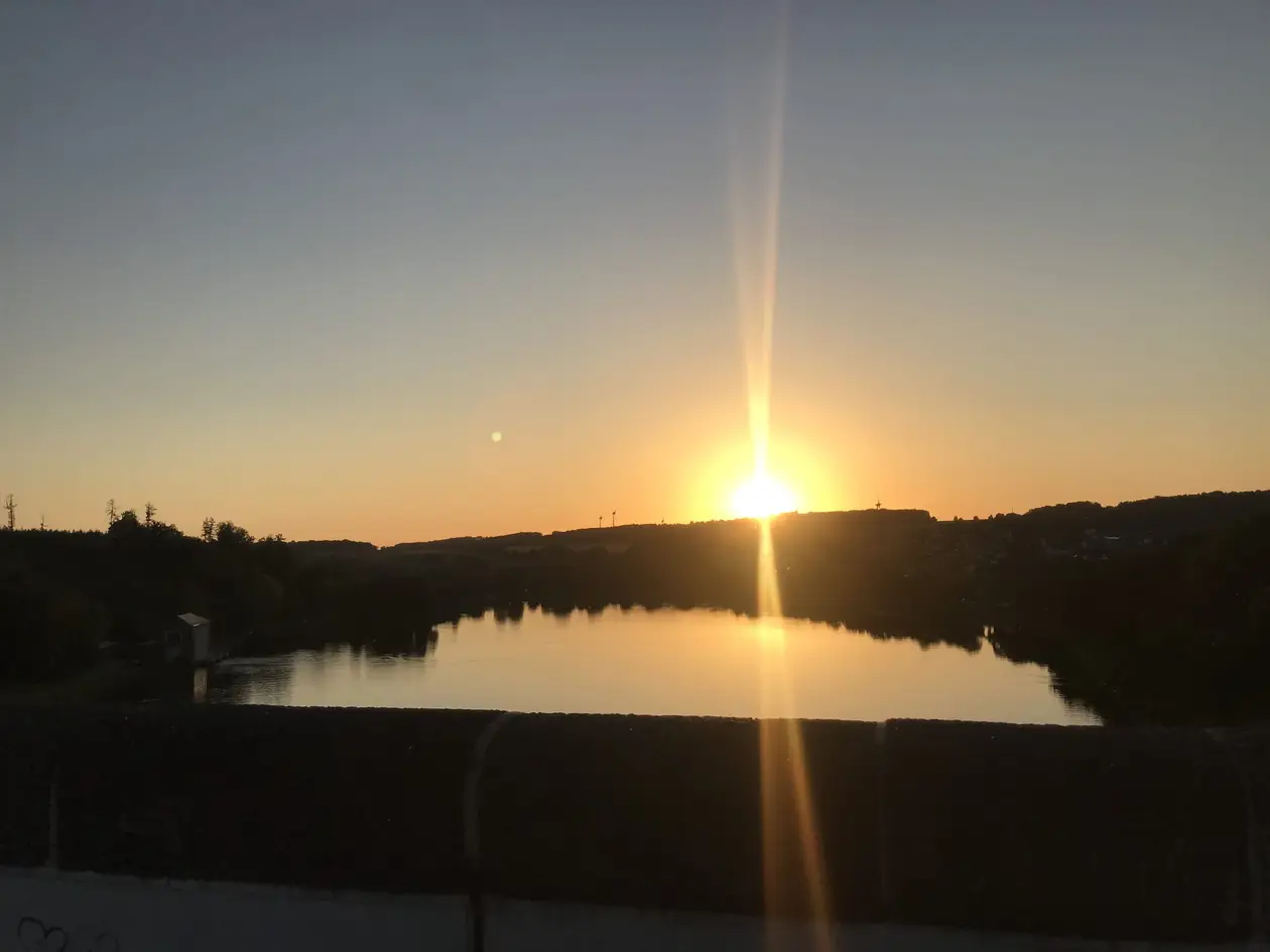 Die Sonne steht knapp über einer Hügelkette und spiegelt sich in einem Gewässer im Tal, vorne die dunkle Brüstung der Staumauer