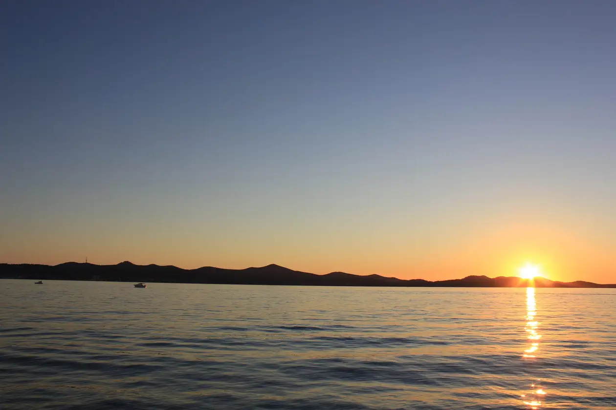Die Sonne steht knapp über der Silhouette einer Insel, davor ruhiges Wasser mit zwei kleinen Booten
