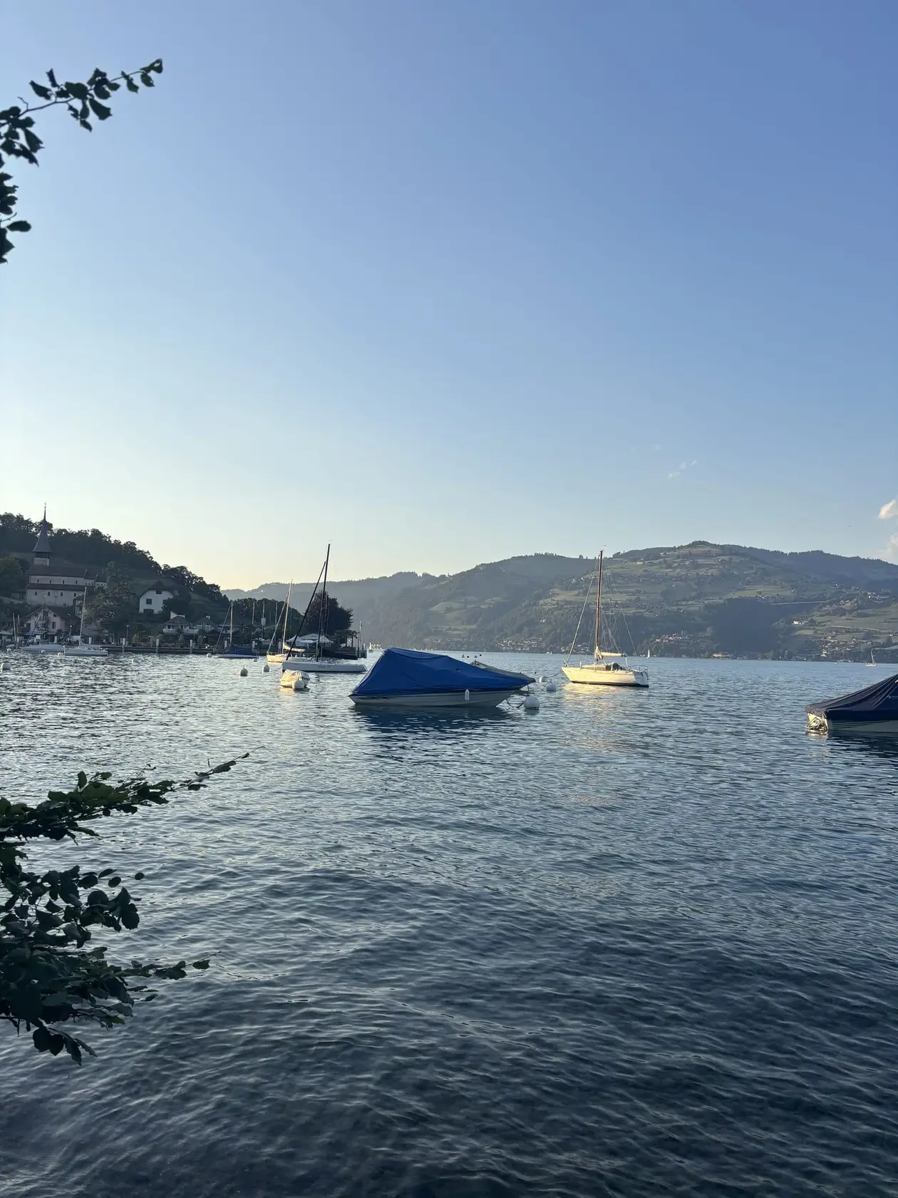 Segelboote und ein abgedecktes Motorboot auf dem See im Abendlicht, am Ufer ein Ort und bewaldete Hügel