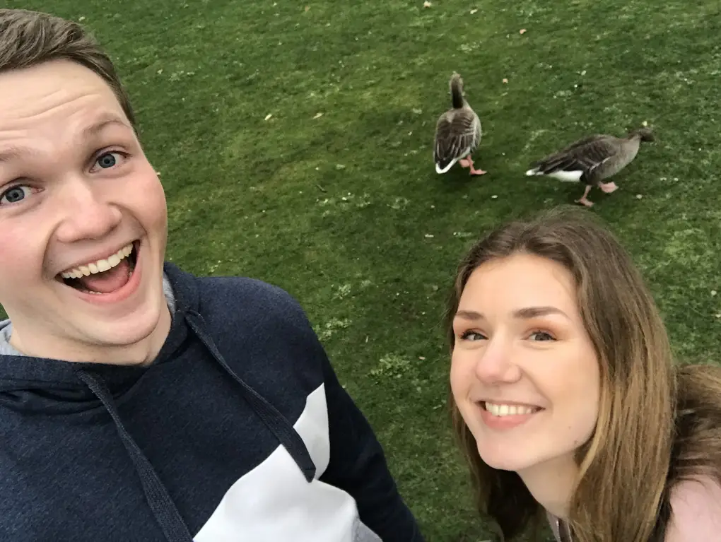 Max grinst mit offenem Mund in die Kamera, neben ihm Rebecca, hinter den beiden zwei Graugänse auf dem Rasen