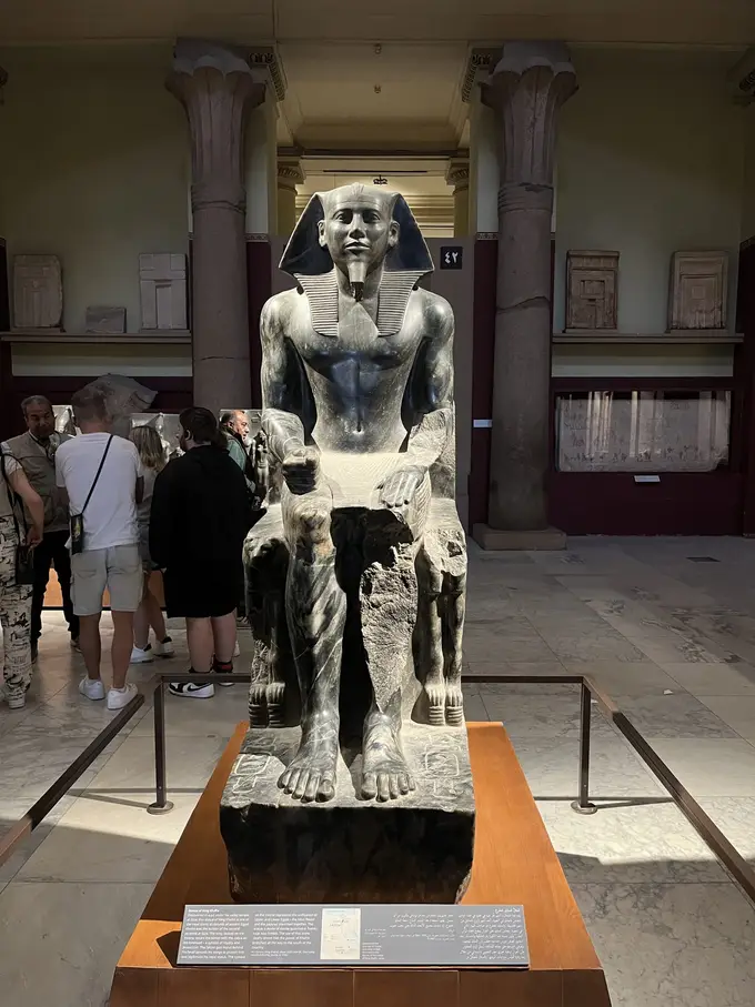 Dunkle Sitzstatue des Königs Chephren mit Kopftuch und Bart auf einem Sockel mit Infotafel, im Hintergrund Säulen und eine Besuchergruppe