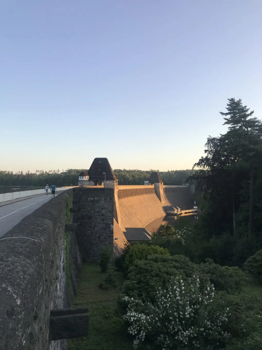 Blick von der Mauerkrone an der Staumauer aus Bruchstein entlang, die in der Abendsonne leuchtet, oben Türme mit spitzen Dächern und zwei Spaziergänger auf dem Weg