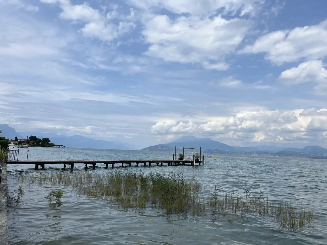 Ein langer Holzsteg führt über den Gardasee zu einer kleinen Plattform mit Sonnensegel, vorne Schilf im Wasser, am Horizont Berge unter Wolken