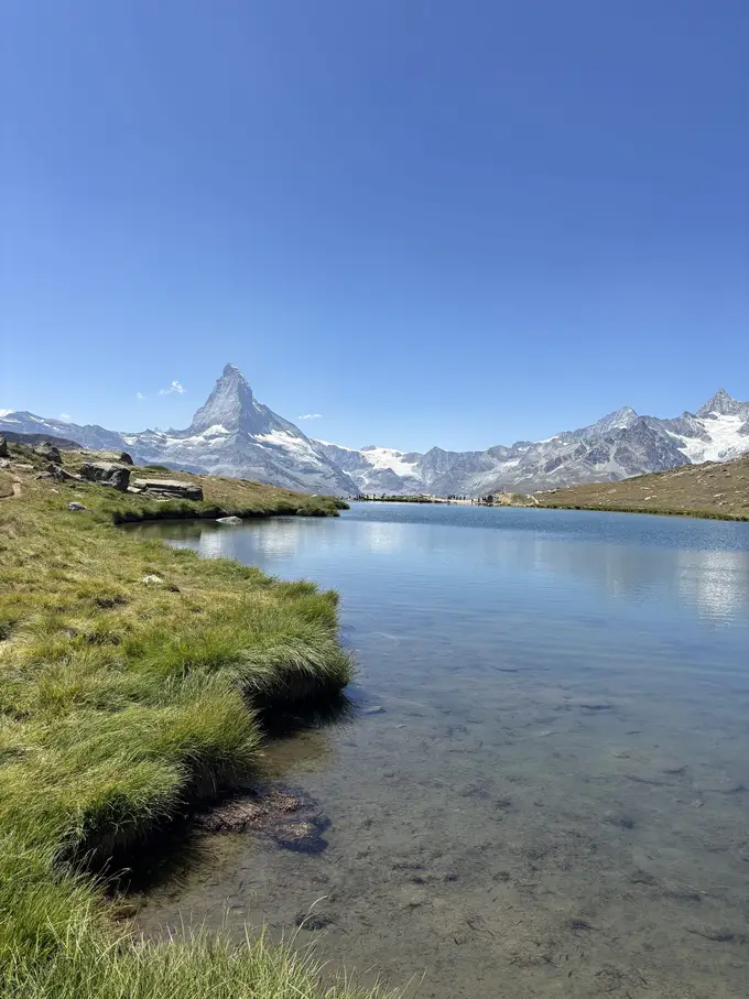 Ein klarer Bergsee mit grasigem Ufer, am Ende das Matterhorn und eine Reihe verschneiter Gipfel unter blauem Himmel