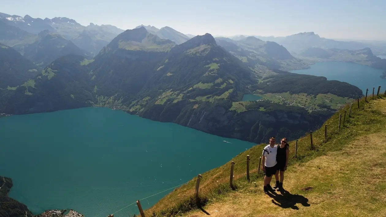 Drohnenfoto: Max und Rebecca stehen auf einem grasigen Grat am Zaun, tief unter ihnen ein türkiser See zwischen steilen Bergen