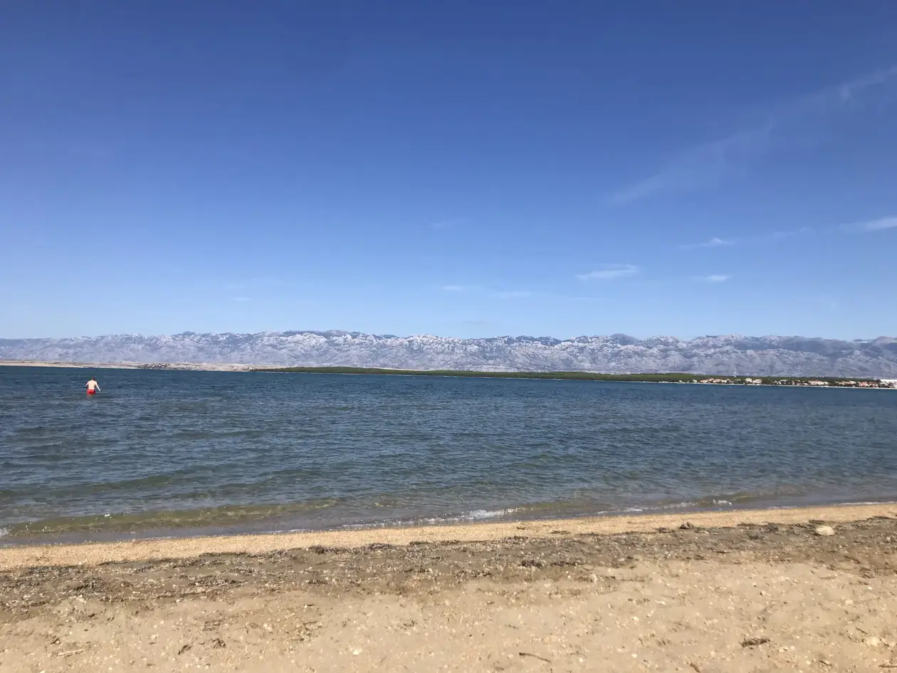 Ein flaches Ufer aus Sand und Kies, im seichten Wasser watet eine Person in roter Badehose, dahinter ein kahler Gebirgszug