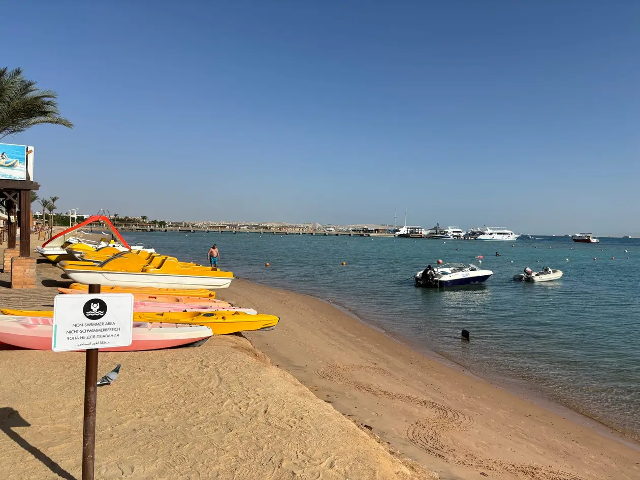 Sandstrand mit aufgereihten Tretbooten und Kajaks, davor ein Schild für den Nichtschwimmerbereich, auf dem Wasser Motorboote und weiter hinten ein Steg mit Ausflugsschiffen