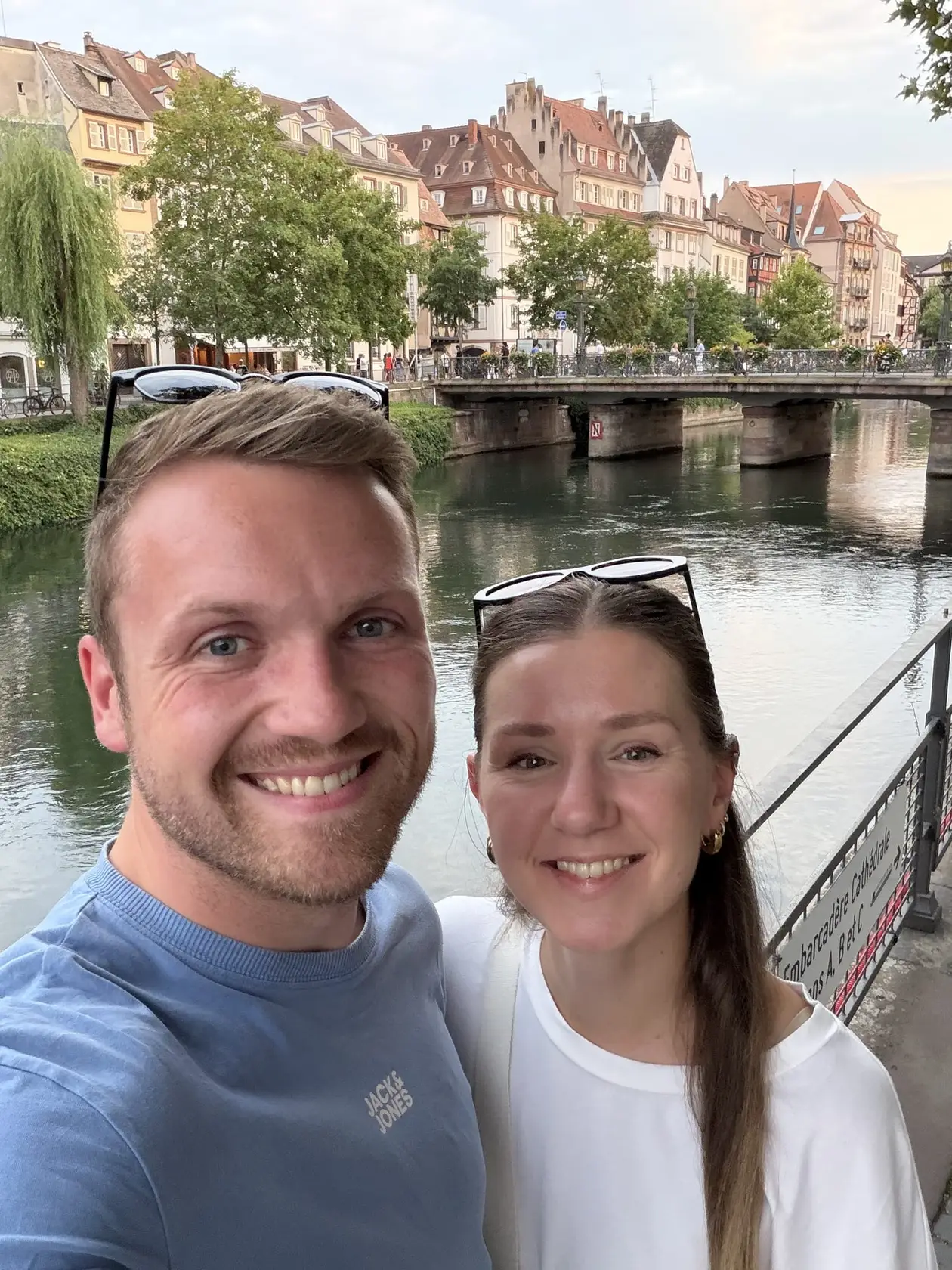 Selfie von Max und Rebecca an einem Fluss, hinter ihnen eine Brücke und eine Häuserzeile im Abendlicht