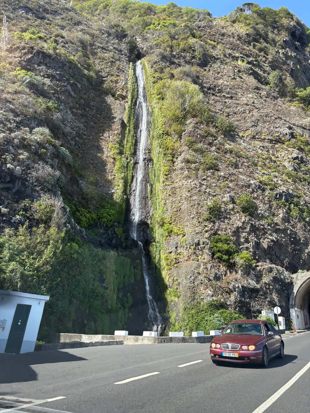 Straße mit Tunneleinfahrt führt an einem Felshang entlang, aus dem ein schmaler Wasserfall herabläuft, ein roter Oldtimer fährt vorbei