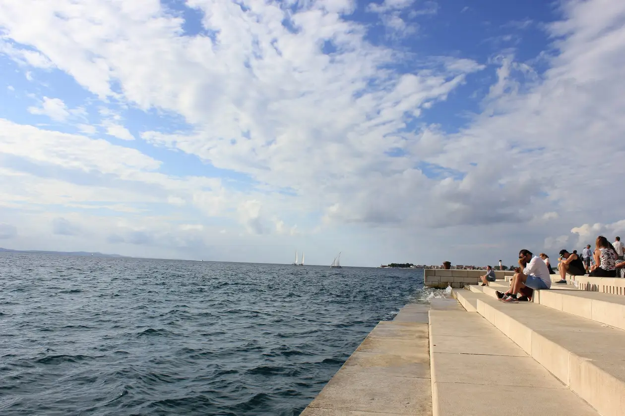 Breite weiße Steinstufen führen ins Meer, eine Welle schwappt über die unteren, rechts sitzen Menschen auf den Stufen, draußen Segelboote