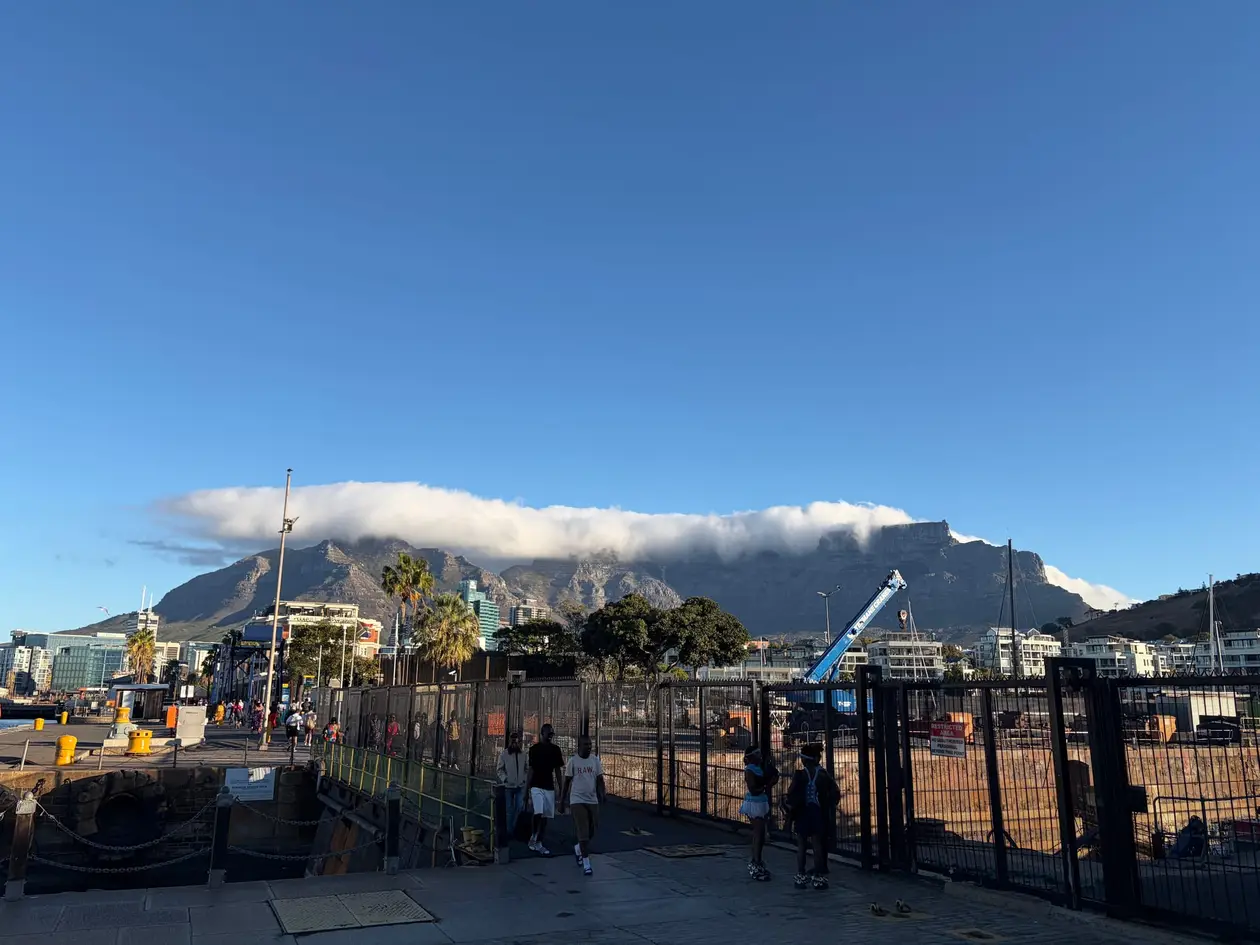 Der Tafelberg mit einer Wolkendecke über dem Hafen der V&A Waterfront, im Vordergrund Kräne und ein Boot