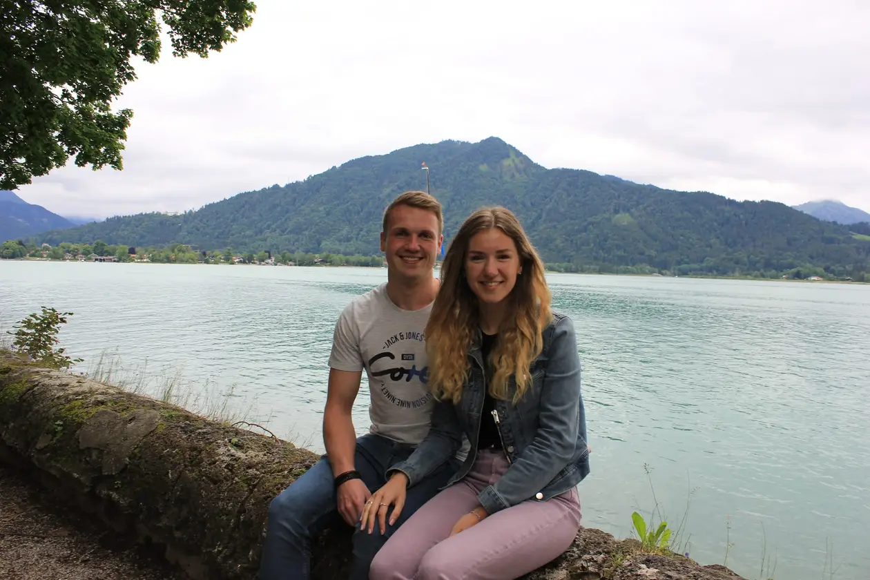 Max und Rebecca sitzen auf einer bemoosten Ufermauer, hinter ihnen der türkisgrüne See und ein bewaldeter Berg unter grauem Himmel