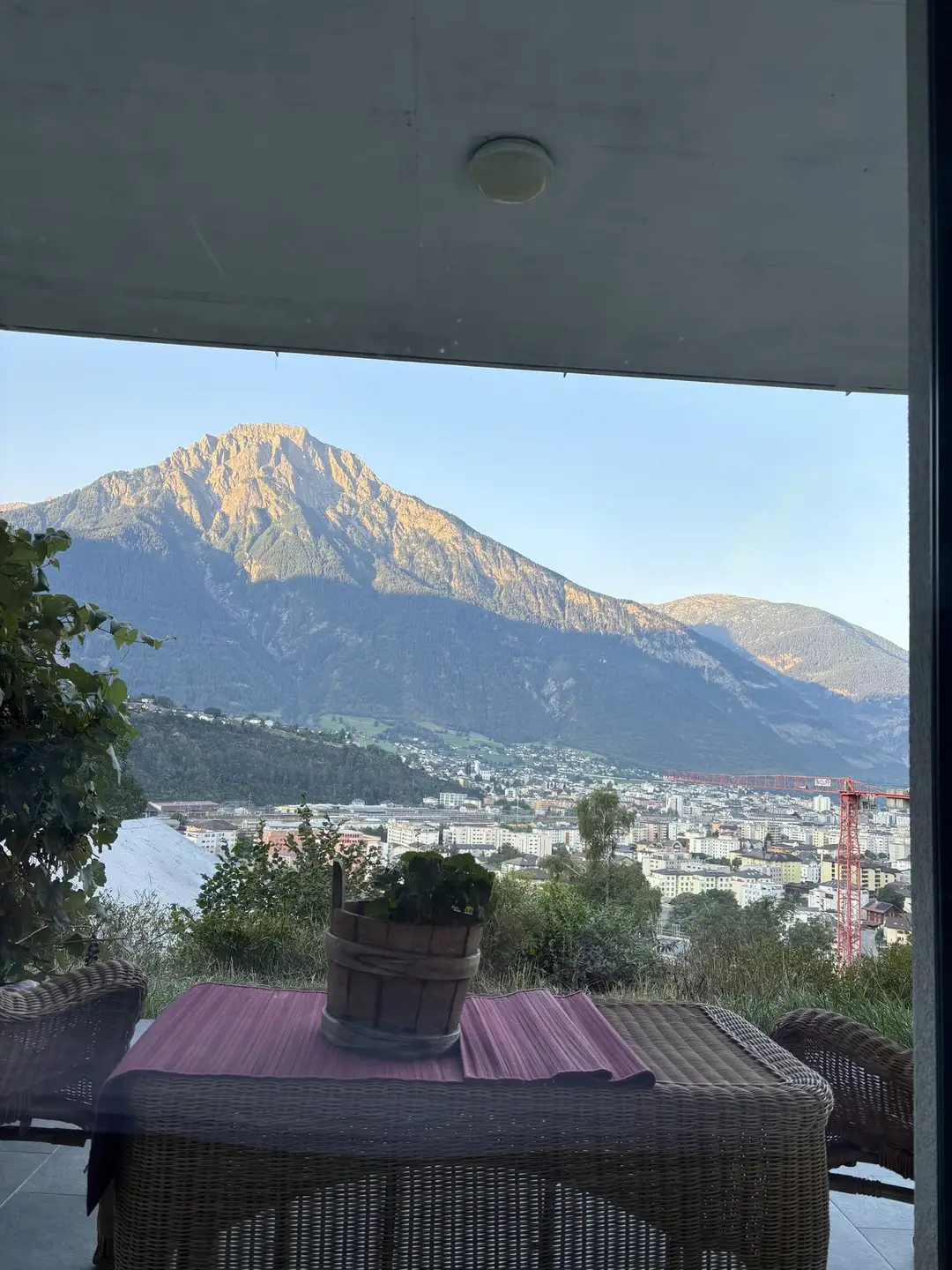Blick von einer überdachten Terrasse mit Rattantisch und Blumentopf auf einen Berg in der Morgensonne, darunter die Häuser einer Stadt im Tal und ein Baukran