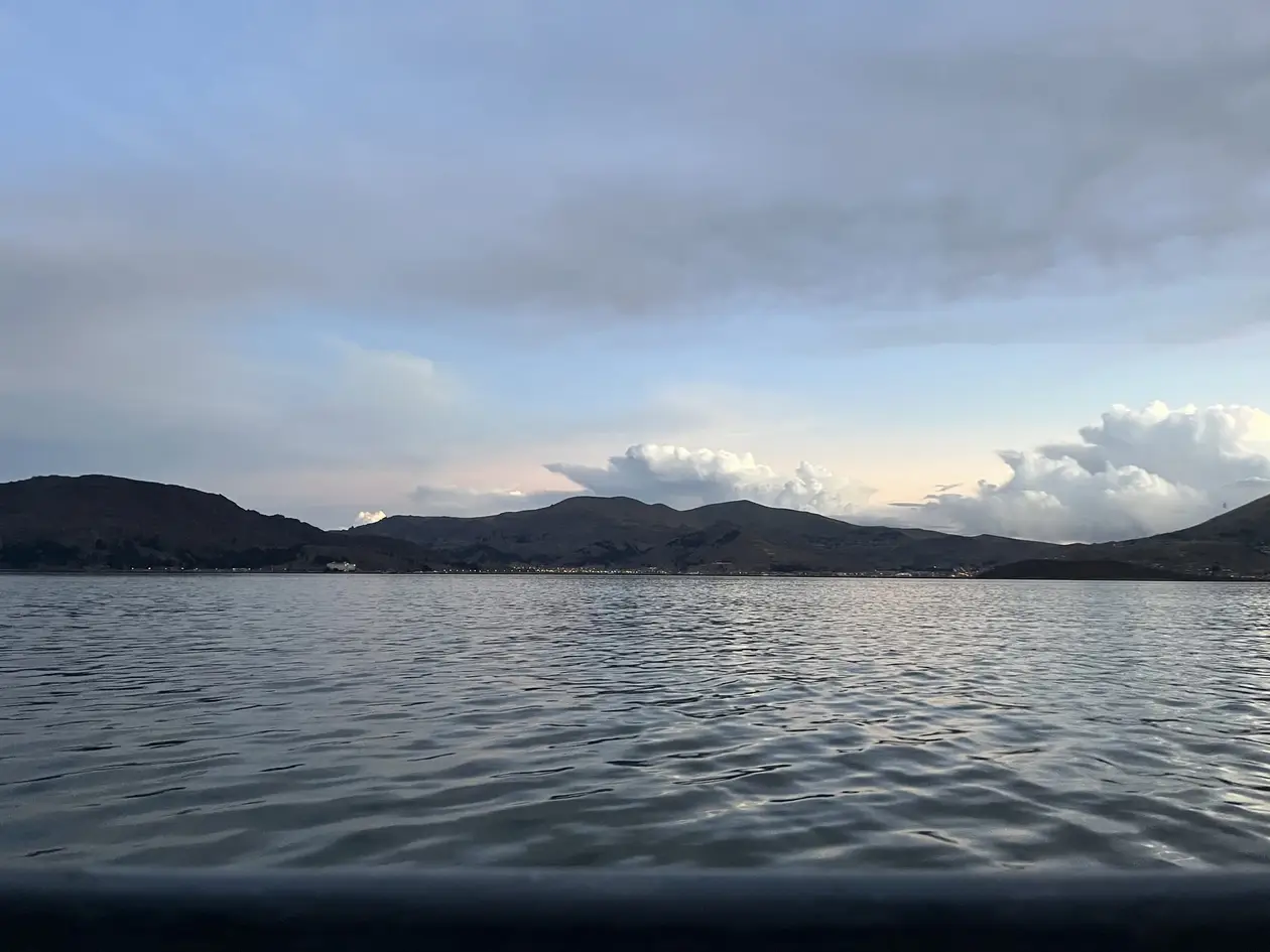 Der Titicacasee unter Wolken, am Ufer dunkle Hügel, vorne die Reling des Bootes