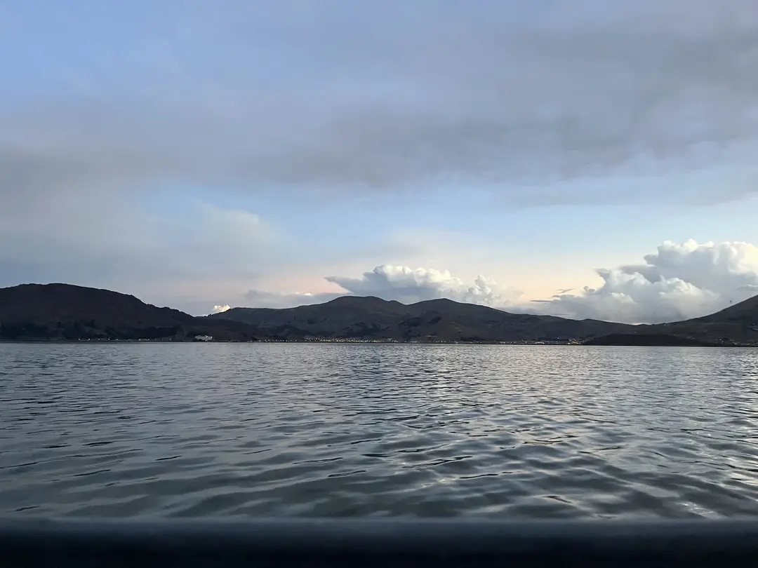 Der Titicacasee unter Wolken, am Ufer dunkle Hügel, vorne die Reling des Bootes