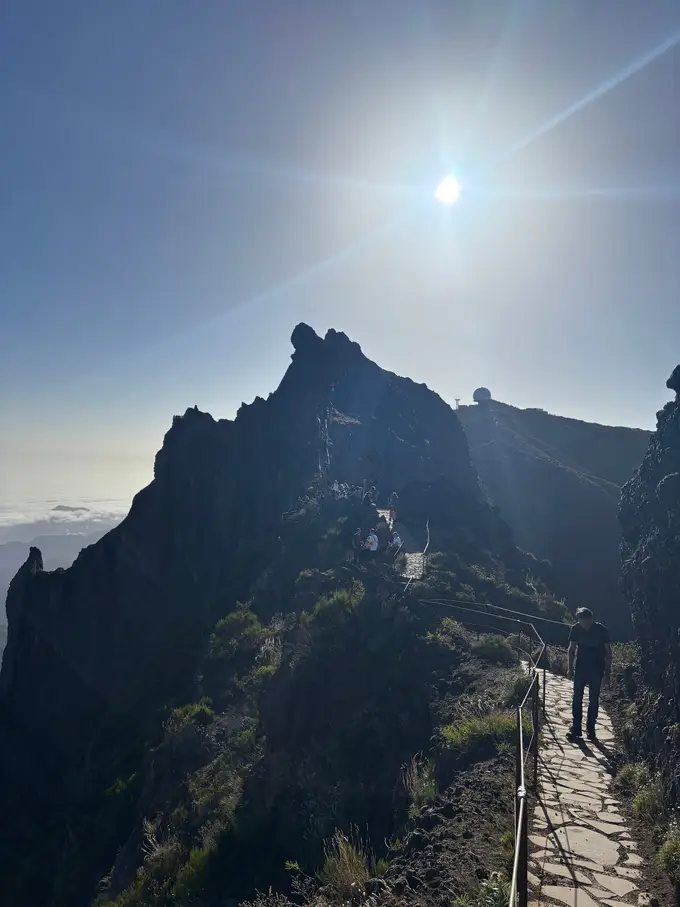 Wanderer steigen eine steinerne Treppe zu einer Felsspitze hinauf, die Sonne blendet von oben