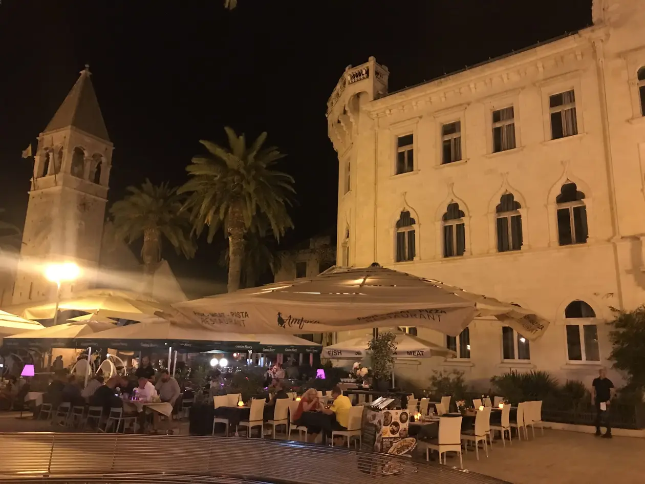 Ein Platz bei Nacht mit Palmen, einem angestrahlten Glockenturm und einem hellen Palast, davor Restaurantschirme und Gäste