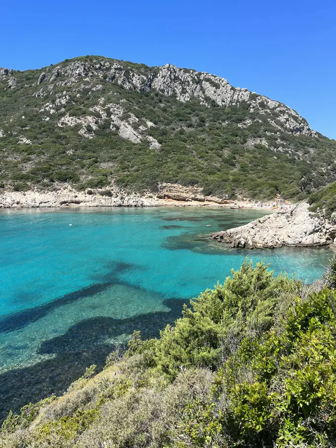Türkise Bucht mit dunklen Felsflecken im Wasser unter einem bewachsenen Berghang