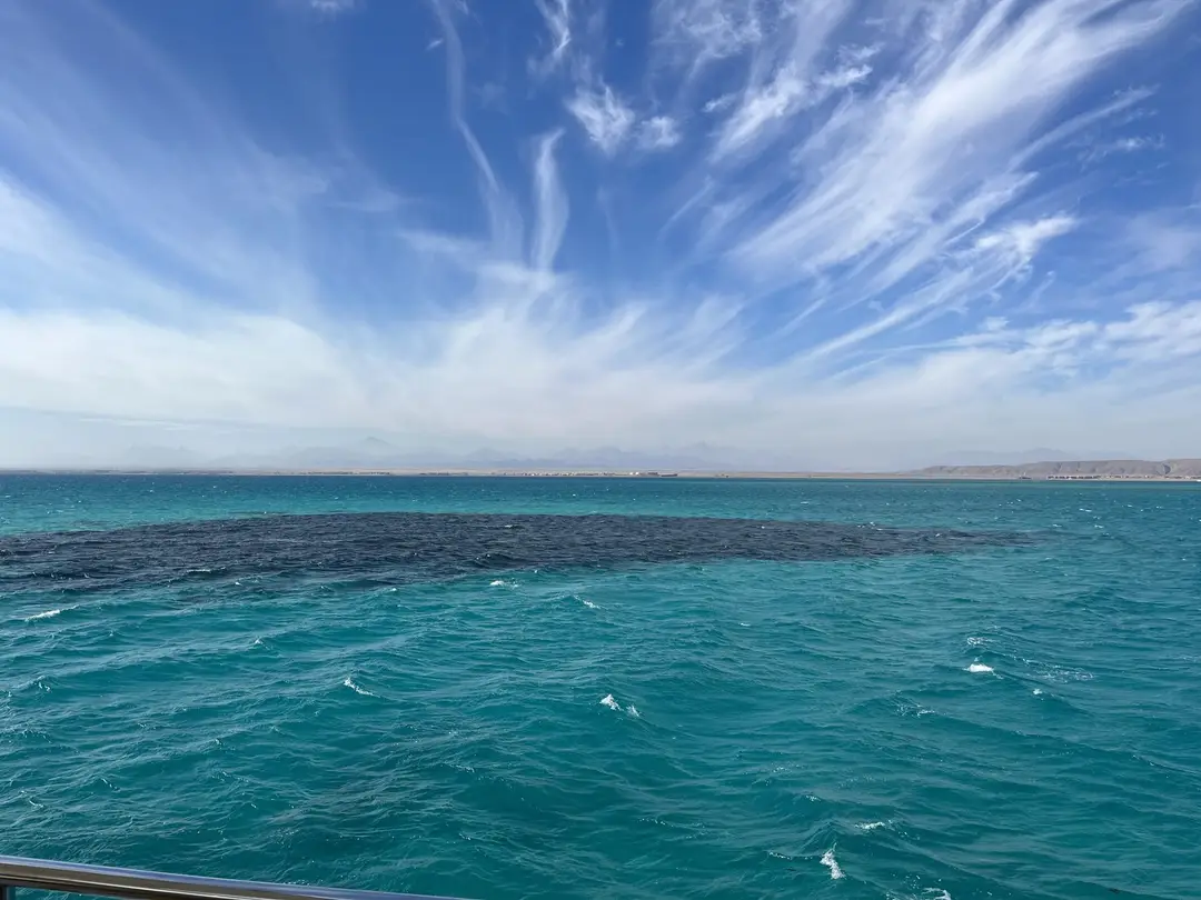 Türkises Meer mit einer großen dunklen Fläche im Wasser, dahinter ein flacher Küstenstreifen und eine Bergkette im Dunst