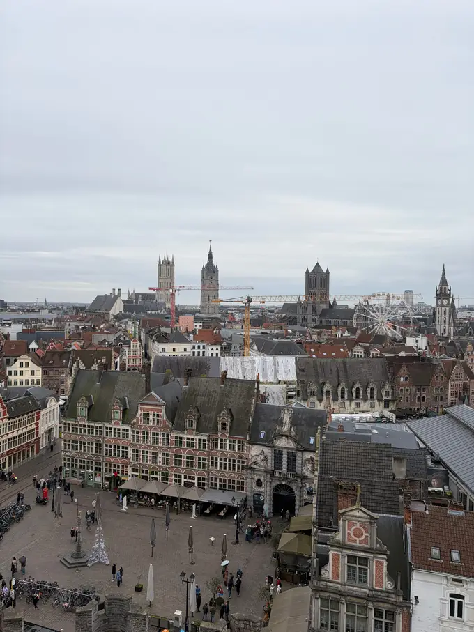 Blick von oben auf einen Platz mit Backsteinhäusern und Terrassen, dahinter die Türme der Genter Altstadt, Baukräne und ein Riesenrad