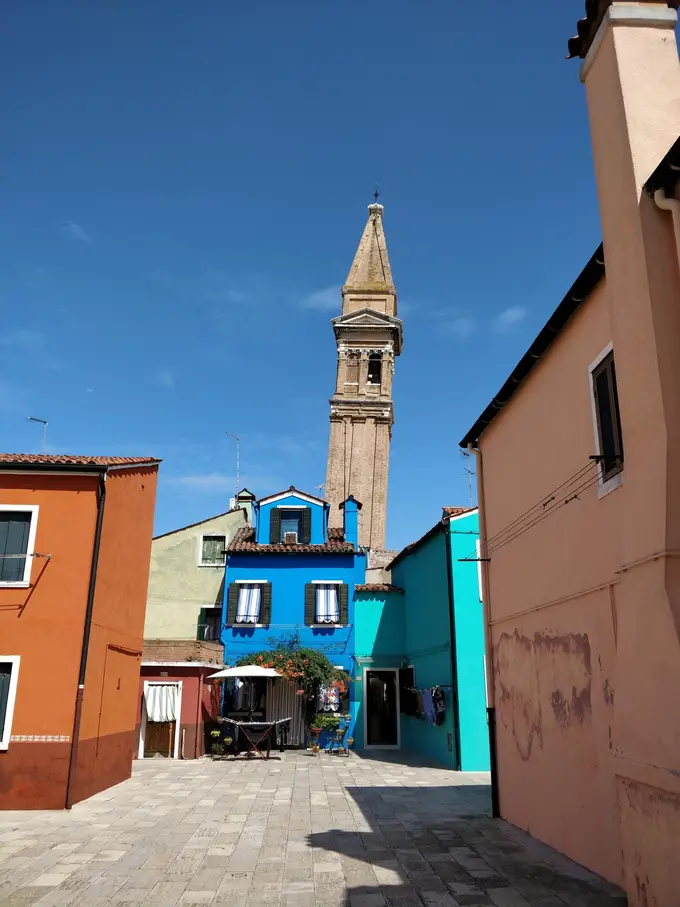 Ein schiefer Glockenturm ragt zwischen einem blauen, einem türkisen und einem orangen Haus in den Himmel