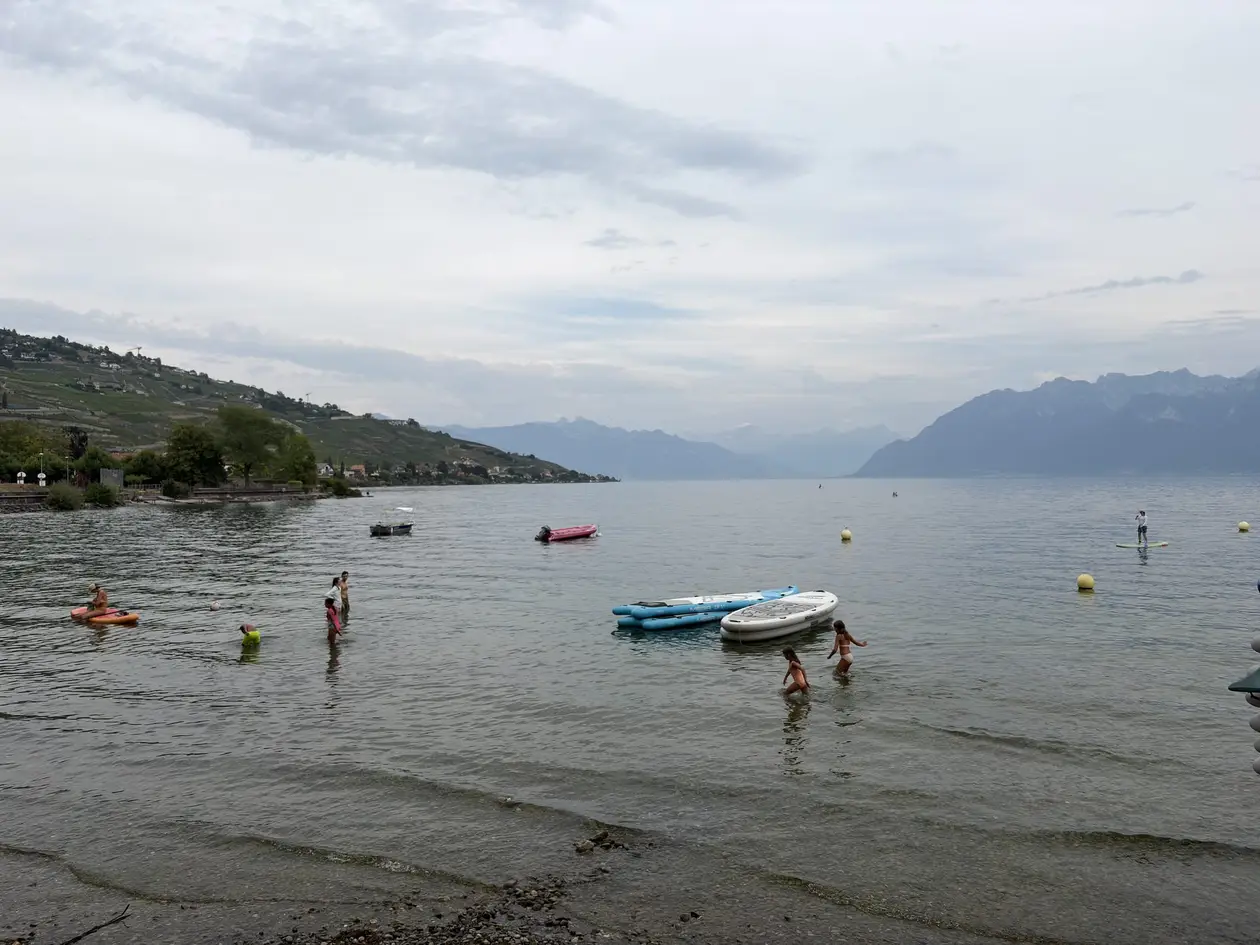 Flaches Ufer am Genfersee mit Badenden, Paddleboards und kleinen Booten, links Weinberge am Hang, in der Ferne Berge