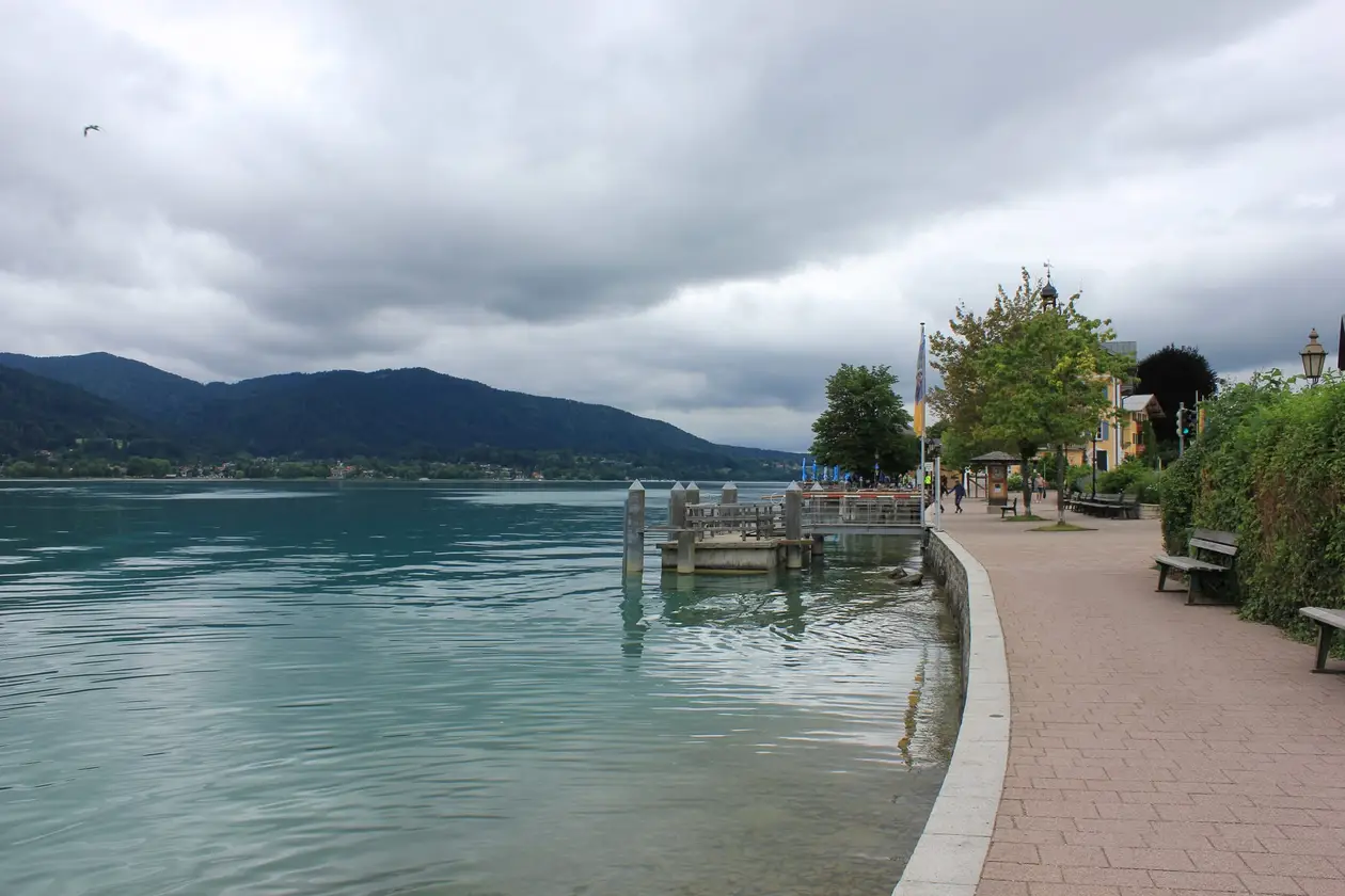 Gepflasterte Uferpromenade mit Bänken und Laternen, links das türkisgrüne Wasser mit einem Steg, dahinter bewaldete Berge unter tief hängenden Wolken