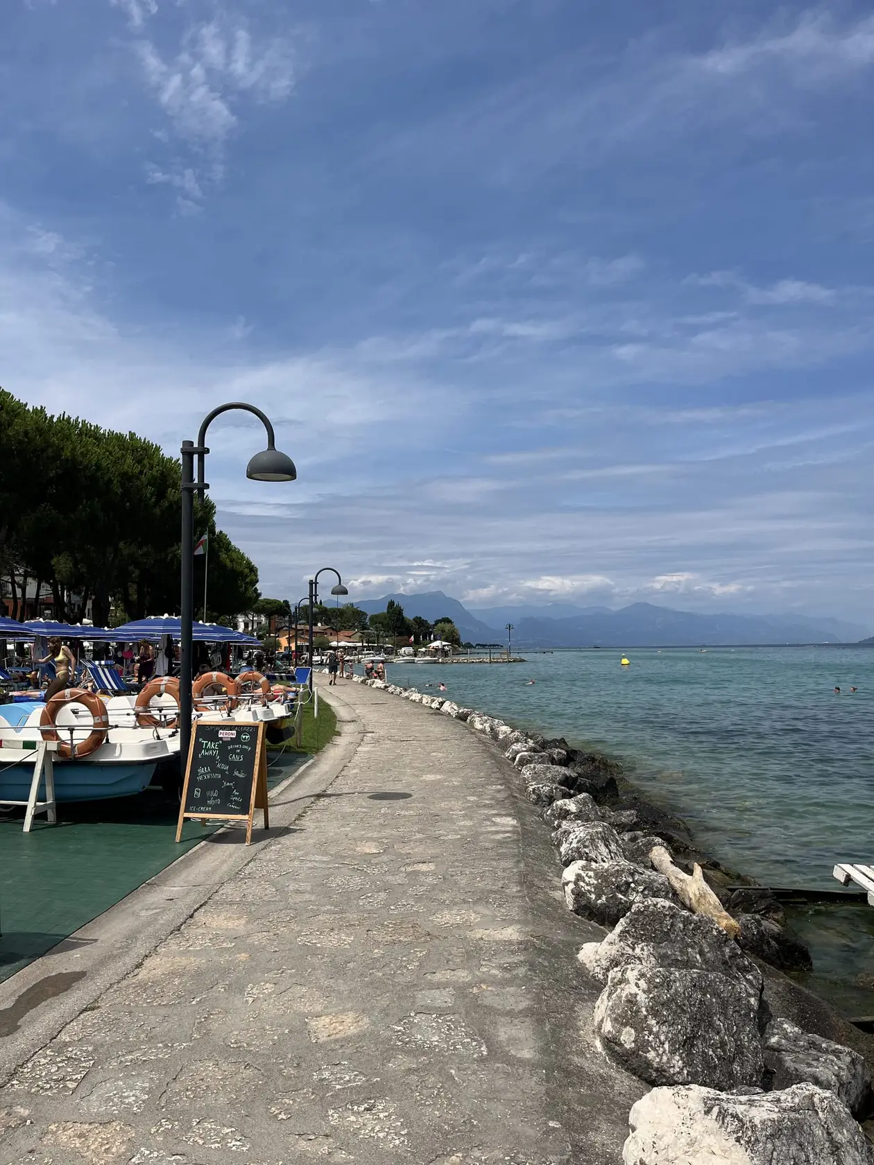 Gepflasterter Uferweg am Gardasee mit Steinblöcken zum Wasser hin, links Tretboote, Sonnenschirme und eine Kreidetafel, am Horizont Berge
