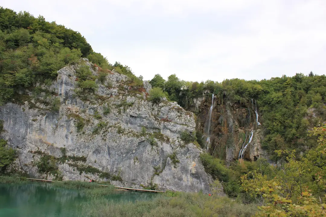 Links eine graue Felswand, rechts fallen dünne Wasserstränge über eine braune Sinterwand, unten ein türkiser See mit Holzsteg