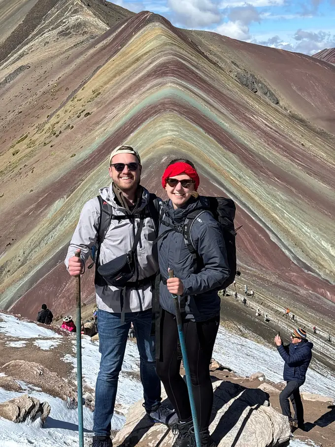 Max und Rebecca mit Wanderstöcken vor den breiten farbigen Streifen des Berges, am Boden liegt Schnee
