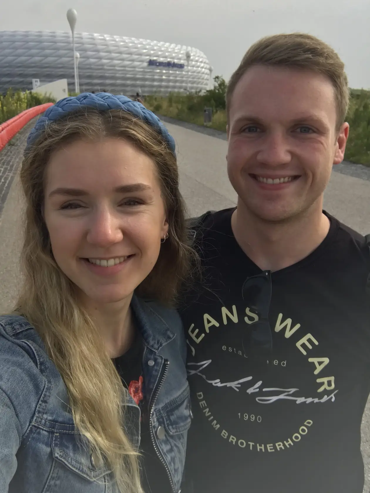 Selfie von Rebecca und Max auf einem breiten Weg, hinter ihnen die weiß schimmernde Fassade der Allianz Arena mit dem Schriftzug