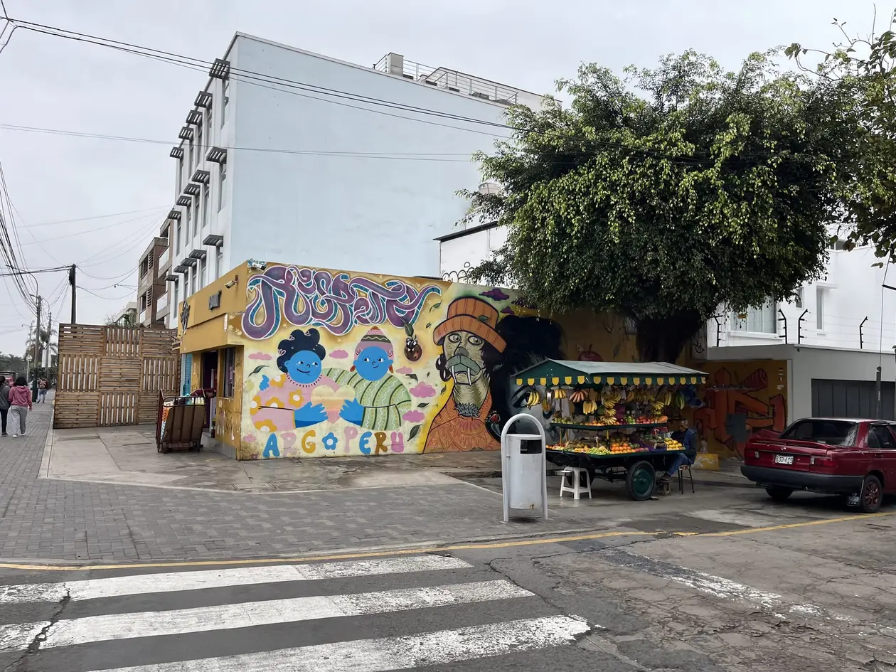 Straßenecke in Barranco, eine gelbe Mauer mit buntem Wandbild und der Aufschrift Arg Peru, davor ein Obststand und ein rotes Auto