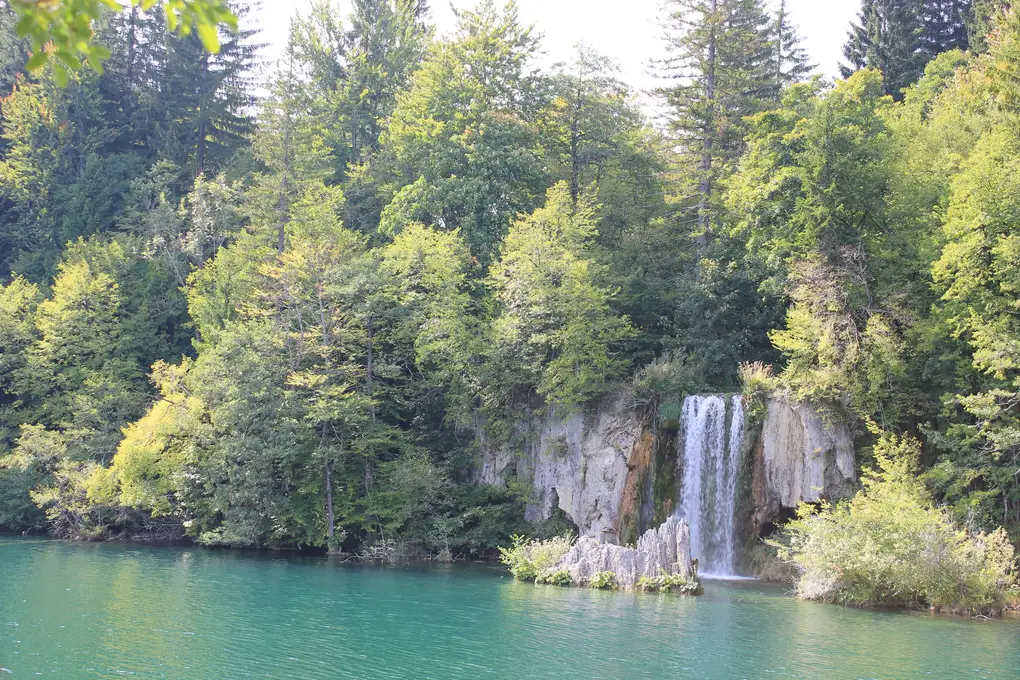 Ein schmaler Wasserfall fällt über eine helle Kalkwand in einen türkisen See, ringsum dichter Wald
